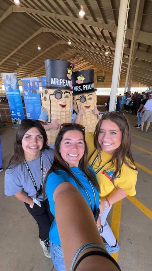 Three young women pose for a selfie with two large peanut mascots at an event.