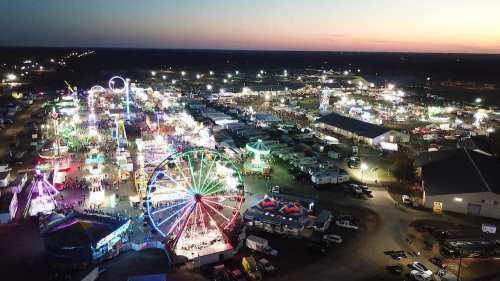 Aerial view of a vibrant fairground at dusk, featuring colorful lights, rides, and bustling crowds.