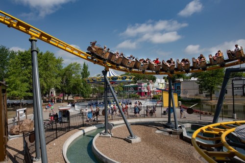 A roller coaster with riders twists above a theme park, surrounded by trees and colorful buildings under a blue sky.