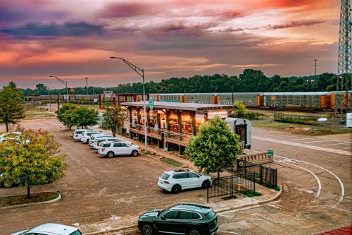 A sunset view of a restaurant with outdoor seating, surrounded by parked cars and a train passing in the background.