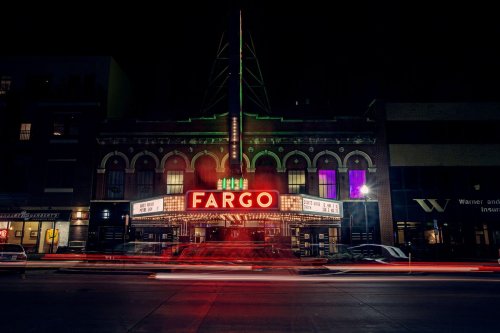 A historic theater at night, illuminated with a bright "FARGO" marquee and colorful lights, with light trails from passing cars.