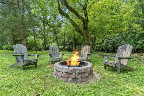 A cozy fire pit surrounded by wooden chairs in a lush green outdoor setting. Flames flicker in the center.