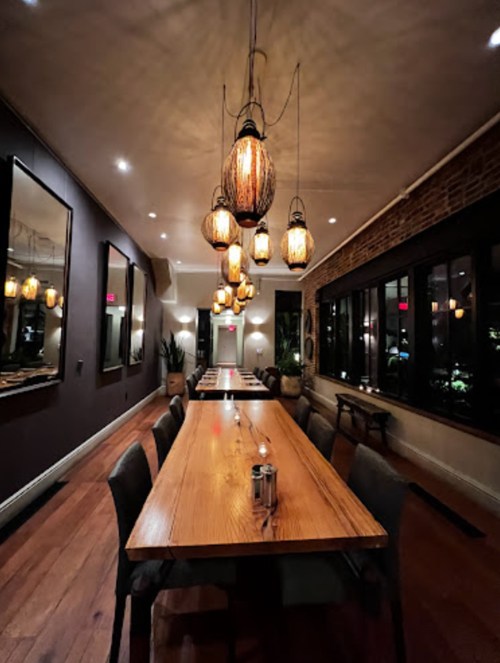 A dimly lit restaurant interior featuring a long wooden table and hanging pendant lights.