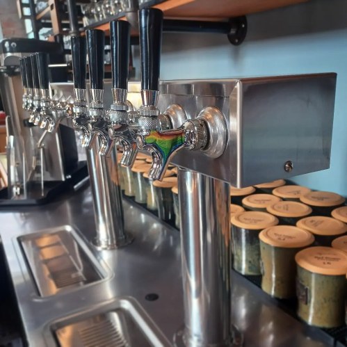 A row of shiny beer taps on a stainless steel bar, with wooden beer kegs in the background.