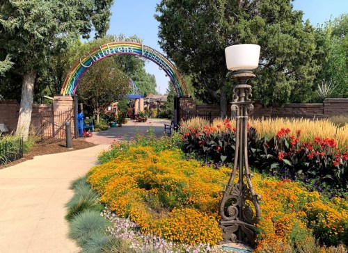 A colorful archway reading "Children's Village" leads into a vibrant garden filled with flowers and greenery.