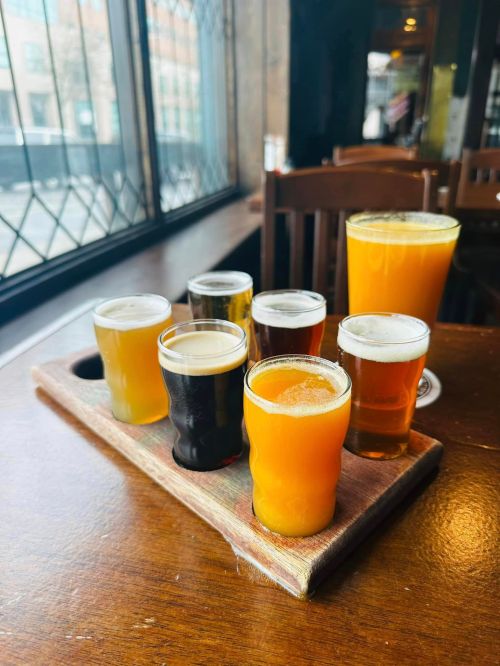 A wooden flight tray holds seven glasses of various beers, with a window view in the background.