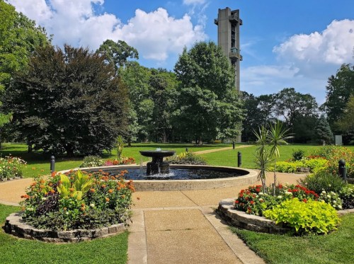 A serene garden with a fountain surrounded by colorful flowers and trees, with a tall tower in the background under a blue sky.