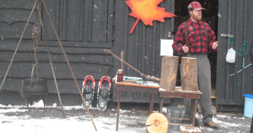 A man in a plaid shirt and suspenders stands outside a cabin, with snowshoes and a pot hanging nearby in a snowy setting.