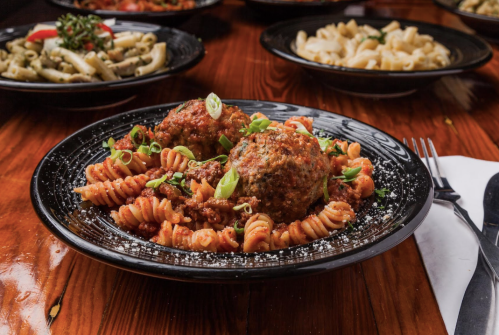 A plate of rotini pasta topped with meatballs and garnished with green onions, surrounded by other pasta dishes.
