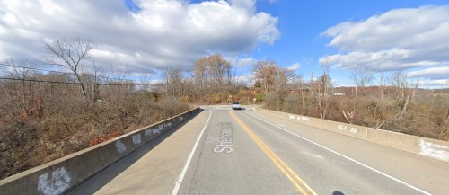 A view of a road bridge with a car, surrounded by trees and a blue sky with clouds.