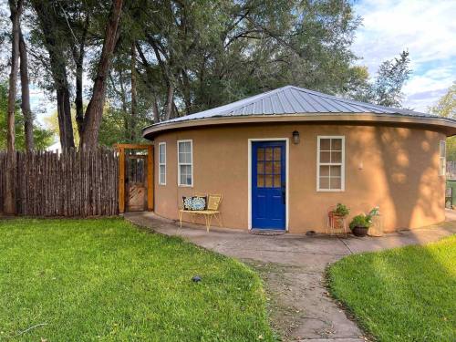 A round, tan house with a blue door, surrounded by green grass and trees, with a wooden fence in the background.