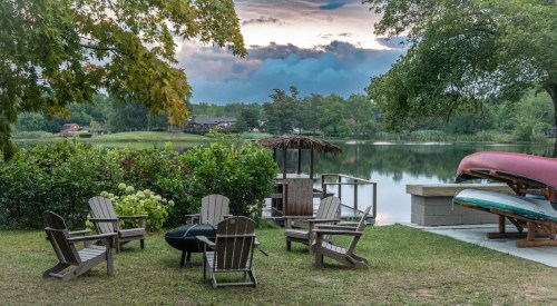 A serene lakeside scene with wooden chairs, a fire pit, and canoes by the water, surrounded by lush greenery.