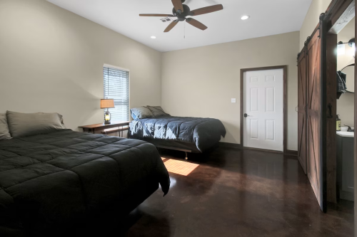 A spacious bedroom with two beds, a lamp, and a door leading to a bathroom, featuring a ceiling fan and natural light.