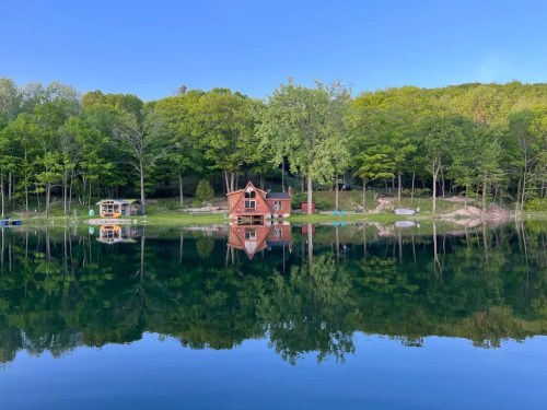 A serene lake scene with a red cabin and lush green trees reflected in the calm water under a clear blue sky.