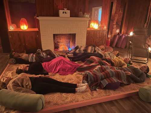 A group of people lying on a soft surface in a cozy room, surrounded by candles and a glowing fireplace.