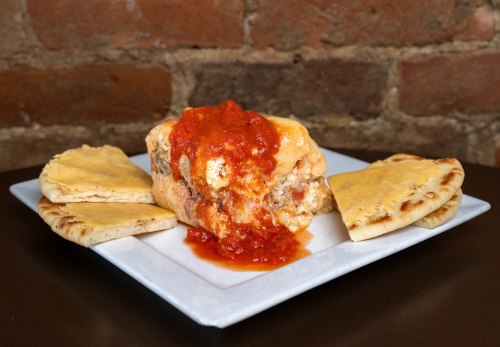 A plate featuring a stuffed dish topped with red sauce, surrounded by pieces of flatbread against a brick background.