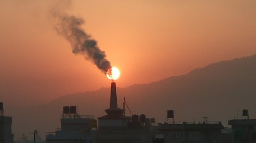 A sunset with a smokestack in silhouette, emitting smoke against a backdrop of mountains and a colorful sky.