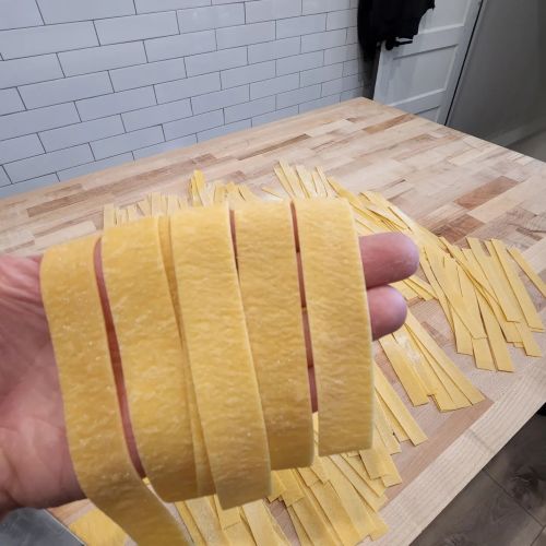 A hand holding fresh, wide strips of pasta over a wooden surface with more pasta laid out in the background.