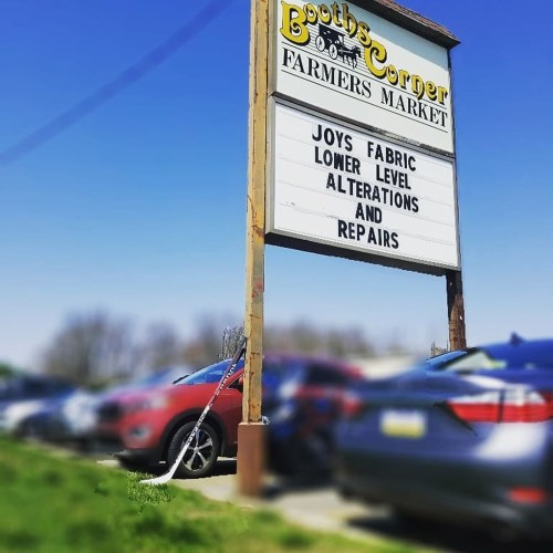 Sign for Booths Corner Farmers Market, advertising Joys Fabric alterations and repairs, with parked cars in the foreground.