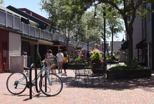 A sunny outdoor plaza with a bike parked, surrounded by greenery and people walking along brick pathways.