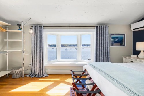 Bright bedroom with a bay window overlooking a serene water view, featuring light curtains and wooden flooring.
