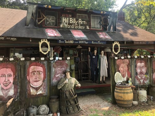 A quirky art gallery with painted portraits, mannequins, and rustic decor, surrounded by trees.