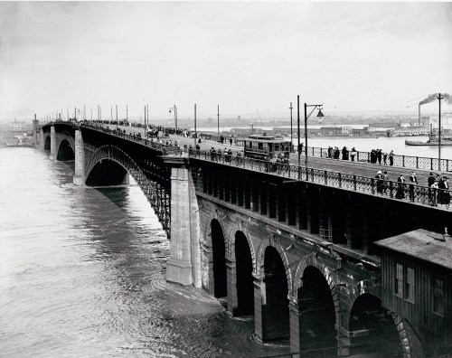 Historic black-and-white photo of a bridge with pedestrians and a tram, spanning a river with boats in the background.