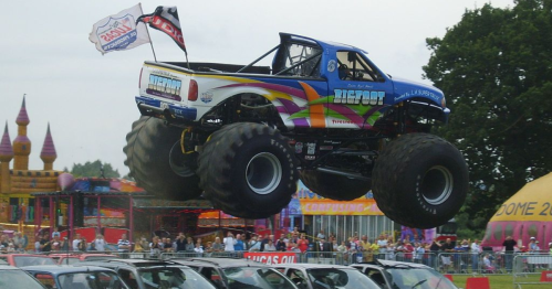 A colorful monster truck jumps over parked cars at a lively outdoor event, with spectators watching in the background.