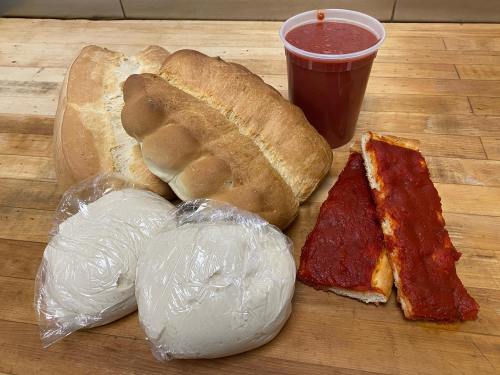 A loaf of bread, two wrapped dough balls, a slice of pizza with sauce, and a cup of marinara sauce on a wooden surface.