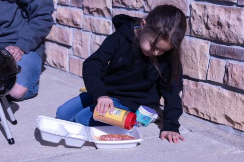 A young girl in a black hoodie pours mustard onto a hot dog in a takeout container, sitting on the ground by a stone wall.