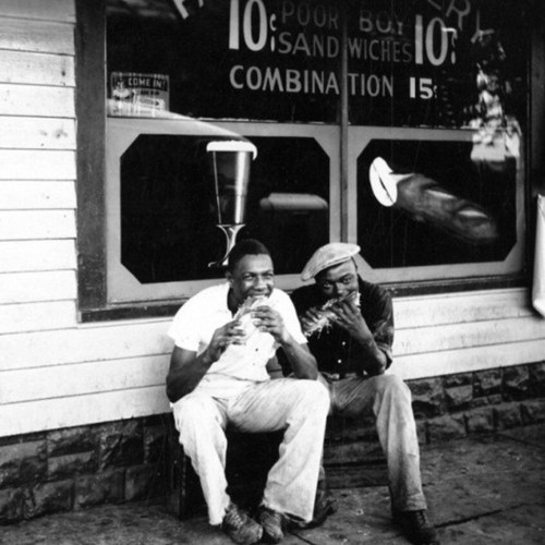 Two men sitting outside a shop, enjoying sandwiches, with a vintage sign in the background advertising food prices.