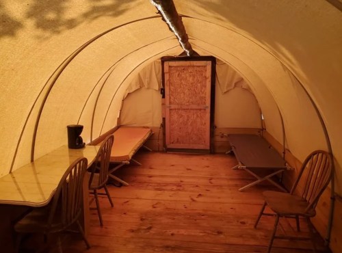 Interior of a tent with wooden floors, two tables, chairs, and a door at the far end. Natural light filters in.