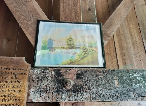 Framed painting of a serene landscape with a river and trees, hanging on a rustic wooden wall.