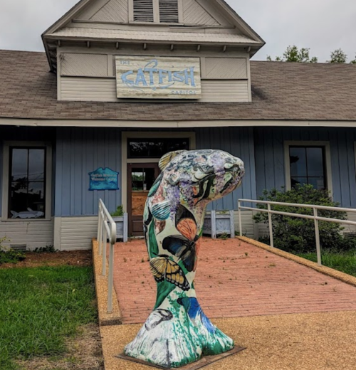 A colorful catfish sculpture stands in front of a restaurant with a sign reading "Catfish Cabin."