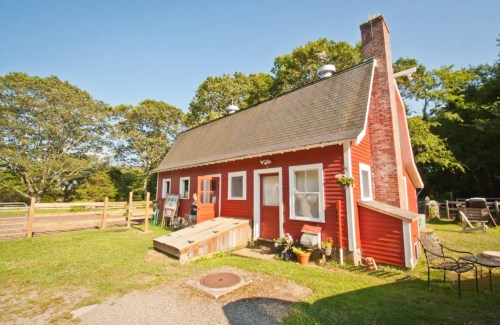 A charming red farmhouse with a sloped roof, surrounded by greenery and a small outdoor seating area.