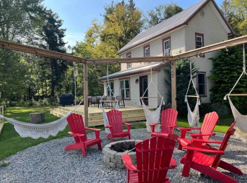 A cozy house with a deck, surrounded by trees, featuring red Adirondack chairs and a fire pit in a gravel area.