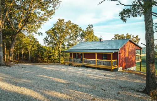 A red cabin with a metal roof sits on a gravel driveway, surrounded by trees and a clear blue sky.