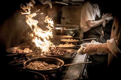 A chef cooking in a busy kitchen, with flames rising from a pan and various dishes being prepared.