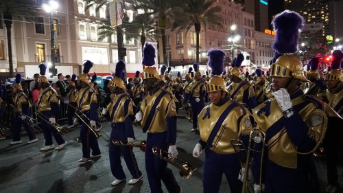 A marching band in blue and gold uniforms parades down a city street at night, with festive lights in the background.