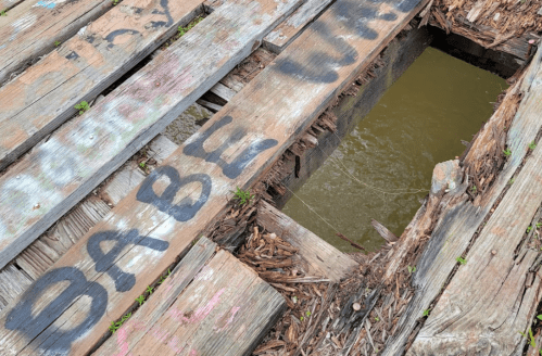 A weathered wooden bridge with a large hole, surrounded by graffiti and exposed wood, over a body of water.