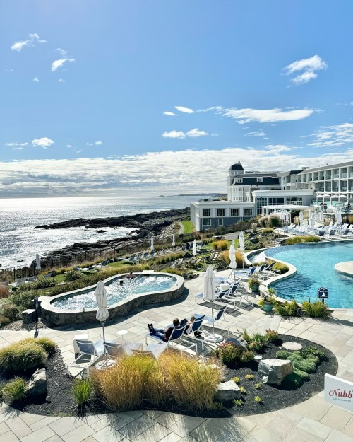 A scenic view of a coastal resort with a pool, hot tub, and lounge chairs overlooking the ocean under a blue sky.