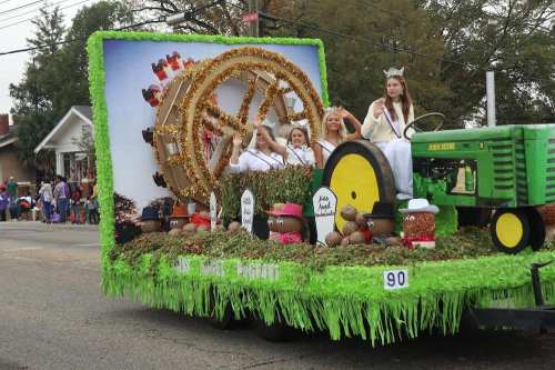 A festive parade float featuring women in crowns, decorated with greenery and a large wheel, celebrating a local event.