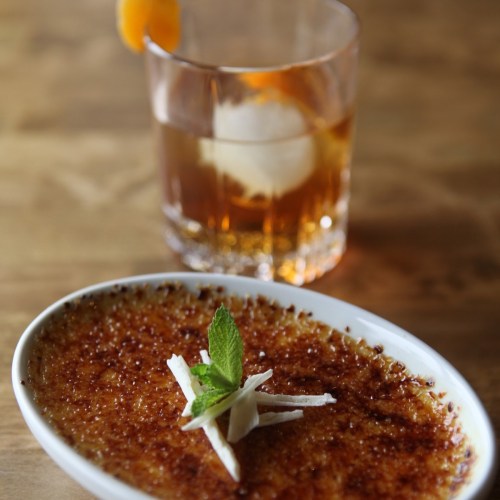 A creamy dessert with a caramelized top, garnished with mint, beside a glass of whiskey with an orange twist.