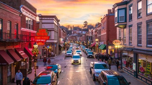 A vibrant street scene at sunset, featuring shops, cars, and people enjoying the lively atmosphere.
