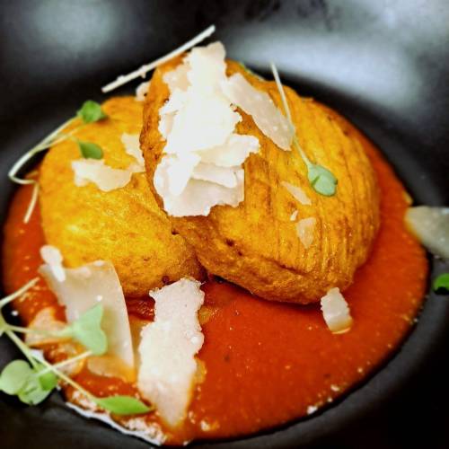 Two golden-brown fritters on a bed of rich red sauce, garnished with shavings of cheese and microgreens.