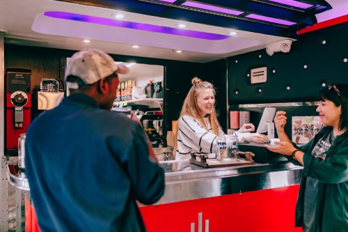 A woman at a food truck counter smiles while serving a customer, with another person waiting nearby.