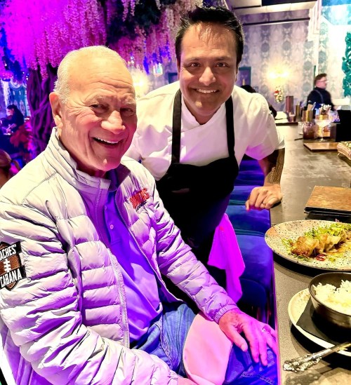 Two smiling men in a restaurant setting, one in a gray jacket and the other in a chef's apron, with food on the table.