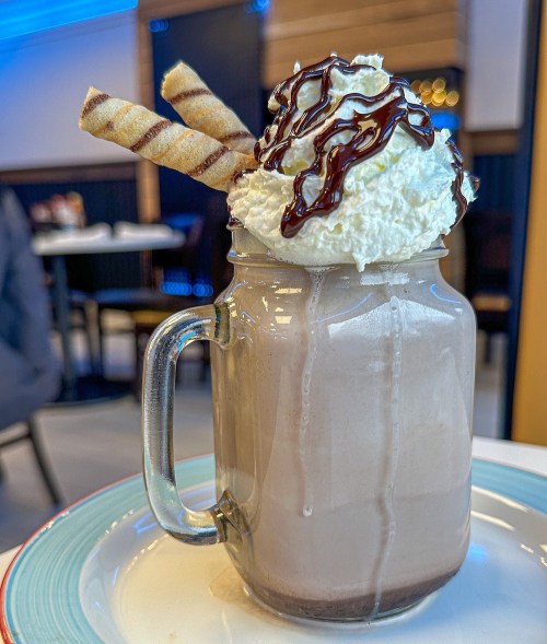 A glass mug filled with chocolate milkshake, topped with whipped cream, chocolate drizzle, and two striped wafer sticks.