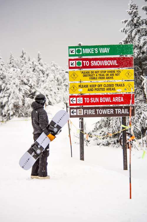 A snowboarder stands near a trail sign in a snowy landscape, holding a snowboard and looking at the directions.