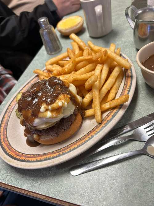 A plate with a burger topped with gravy and mashed potatoes, served with fries and a side of brown sauce.